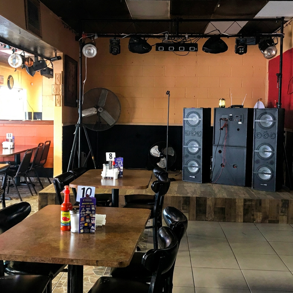 Restaurant dining area with tables, black chairs, bar seating, televisions, and a small stage equipped with speakers and lighting.