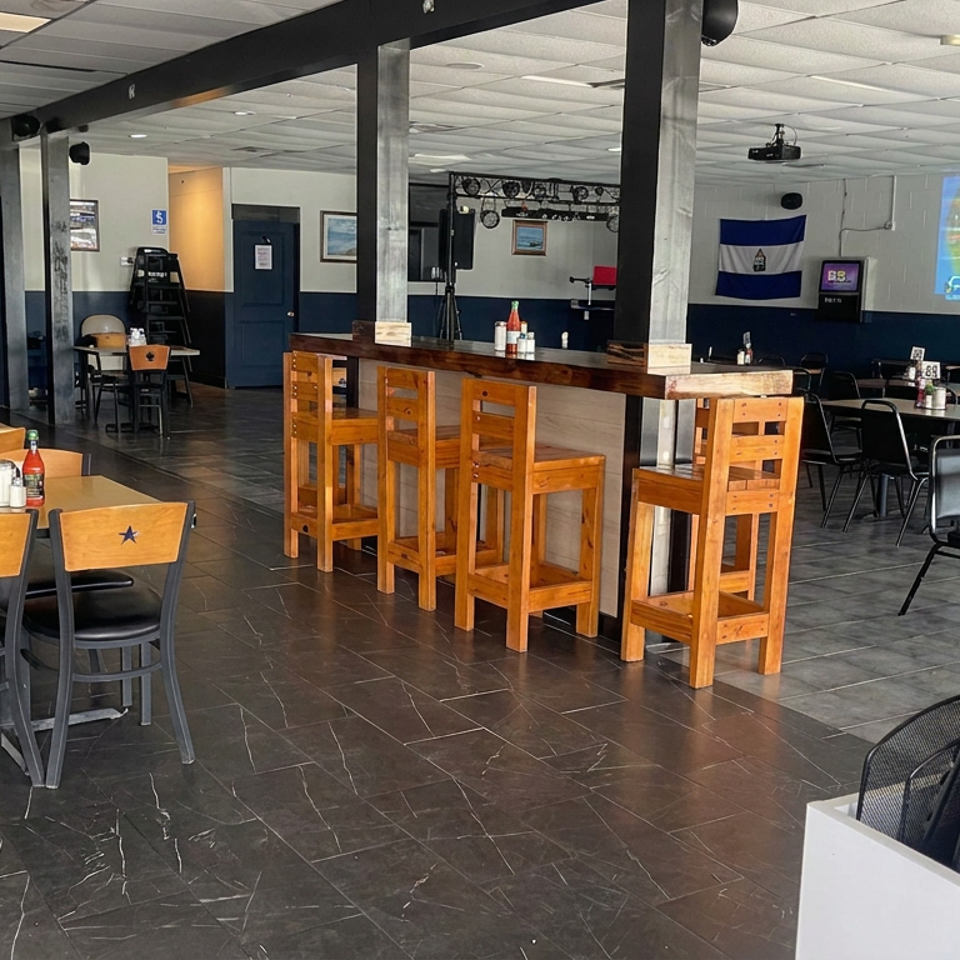 Spacious restaurant dining room with wooden tables, bar seating, condiments, sports memorabilia, and a soccer match projected on the wall.